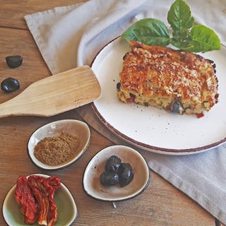 ein Stück Zucchini-Frittata, daneben schwarze Oliven, Knoblauch, getrocknete Tomaten in kleinen Schälchen und ein Holzlöffel 