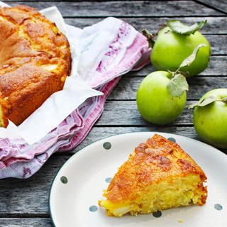 Stück Rührteigkuchen mit Äpfeln auf Teller, daneben ein angeschnittener Kuchen in Backform und Äpfel