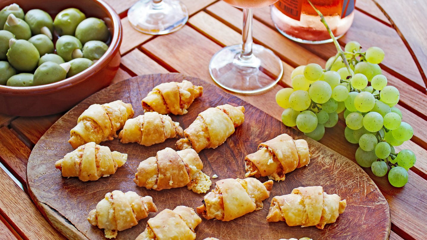Holzbrett mit Käsehörnchen, daneben Trauben und ein Glas Rose-Wein