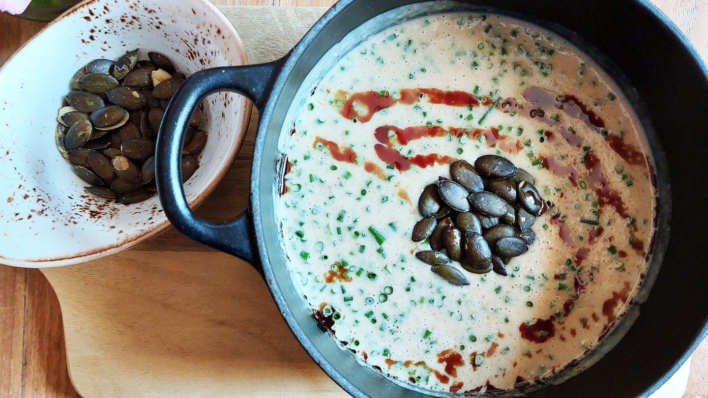 gußeiserner Topf mit heller Suppe mit Kürbiskernen, daneben eine kleine Schüssel mit Kürbiskernen