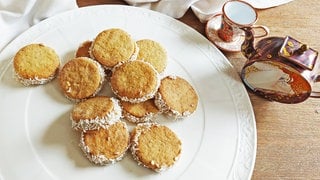 weißer Teller mit hellen Plätzchen mit Kokosraspeln am Rand. Daneben eine Schmuck-Teekanne mit Tasse