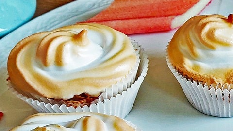 Rhabarber-Muffins mit Baiserhaube auf weißem Teller. Drumherum Stücke von Rhabarber.