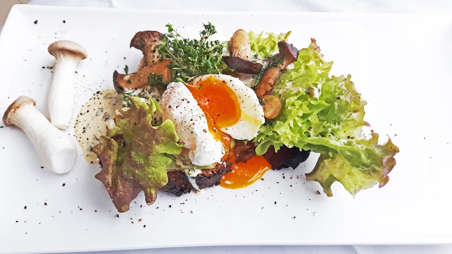 dunkles Brot mit Salat, Pilzen und einem weich gekochten Ei belegt. Daneben zwei Pilze auf dem weißen Teller.