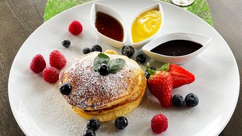 Stapel Pancakes auf einem weißen Teller mit Beeren drumherum, daneben Schälchen mit verschiedenen Saucen, neben dem Teller ein Glas Eierlikör