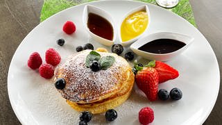 Stapel Pancakes auf einem weißen Teller mit Beeren drumherum, daneben Schälchen mit verschiedenen Saucen, neben dem Teller ein Glas Eierlikör