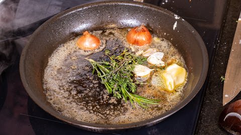 Zum Rezept gehört geschmolzene Butter in einer Pfanne mit Kräutern und Gewürzen, die zum Übergießen der gebratenen Fleischscheiben vom Rind vorbereitet wird.