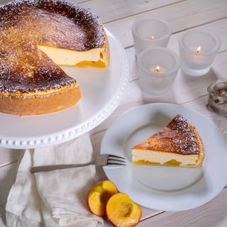 Ein Käsekuchen mit Pfirsich auf einer Kuchenplatte, ein Stück davor angeschnitten auf einem Teller: Bei diesem Rezept verbindet sich eine leckere Füllung mit Quark und ein feiner Boden aus Mürbeteig.