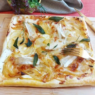 Zwiebelkuchen mit Blätterteig gebacken, darauf Ziegenkäse und Salbei. Der Kuchen liegt auf einem Holzbrett mit einer goldenen Gabel in der Mitte