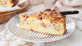 Rezept Zwiebelkuchen badischer Art mit Speck und Kümmel: Ein Stück Zwiebelkuchen liegt zusammen mit Besteck auf einem gestreiften Teller, im Hintergrund ist unscharf weiterer Zwiebelkuchen zu sehen.