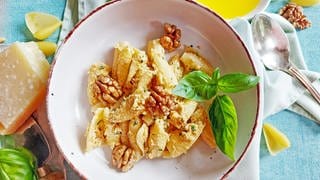 Rezept für Walnuss-Pesto: Nudeln mit Walnusspesto und ganzen Walnüssen und etwas Basilikum in einer hellen Schale, daneben ein Stück Parmesan, eine Serviette und ein silbener Löffel 