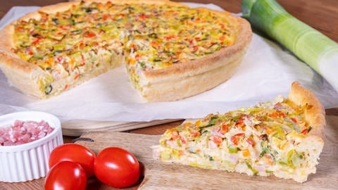 Rezept Lauchkuchen: Der Lauchkuchen ist angerichtet auf einem Holzbrett. Daneben liegt ein Lauch, kleine Tomaten und ein Schälchen mit Schinkenwürfeln.