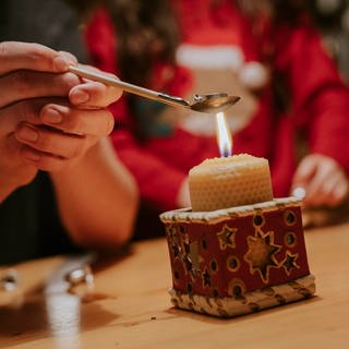 Bleigiessen an Silvester - zwei Hände halten einen Löffel mit Holzstiel, auf dem sich ein Stück Blei befindet, über einer Honigwachskerze