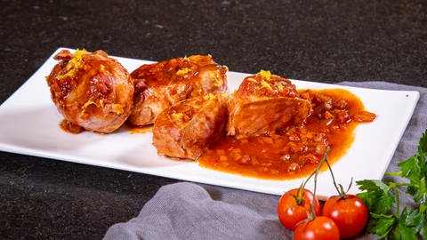 Schweinemedaillons im Speckmantel mit viel Sosse auf einem Teller angerichtet. Im Vordergrund liegen frische Tomaten.
