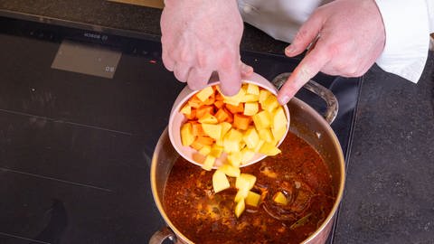 Der Koch Jörg Ilzhöfer kippt in den Topf, mit dem ungarischen Gulasch eine Schüssel kleingeschnittene Karotten- und Kartoffelwürfel.