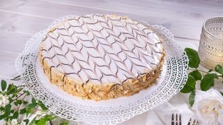 Die Esterhazy-Torte steht nach dem Backen, wie im Rezept beschrieben, schön verziert auf einer Tortenplatte auf einem festlich gedeckten Tisch. Sie schmeckt wie das Original. 