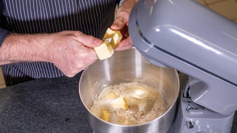 Zwei Hände bereiten an einer grauen Teigrührmaschine einen Brezelteig zu und schneiden Butter in die Masse.