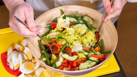 Wie im Rezept beschrieben, werden die Zutaten des Salates in einer Schüssel vermengt. Der Nizzasalat besteht unter anderem aus Ei, Thunfisch und Oliven. 