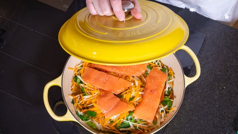 Roher Lachs mit frischem Gemüse in einer Pfanne vor dem Dünsten. Die Pfanne wird mit einem Deckel verschlossen.