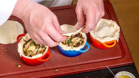 Ein Koch befüllt keine Pastetenformen mit einer Pilzmischung und legt kleine Teigdeckel oben drauf.