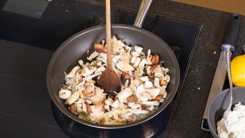 Kleingeschnittene Pilze und Zwiebeln werden in einer Pfanne angebraten und mit einem Holzlöffel umgerührt.