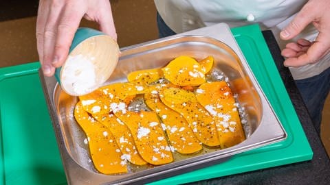 Die Scheiben des Butternut-Kürbis werden für das Rezept mit den Linsen in einer Schale mit Olivenöl beträufelt und mit Puderzucker bestäubt.