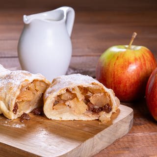 Rezept für Apfelstrudel: Leckerer Apfelstrudel bestäubt mit Puderzucker und Vanillesoße. Der Apfelstrudel ist mit zwei Äpfeln und einem Kännchen mit Vanillesoße angerichtet. 