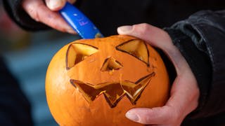 Halloween: Jemand hält einen Kürbis mit Gesicht und ein Messer zum Schnitzen in den Händen