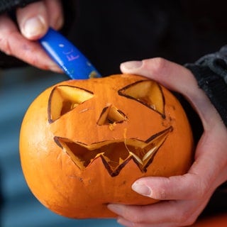 Halloween: Jemand hält einen Kürbis mit Gesicht und ein Messer zum Schnitzen in den Händen