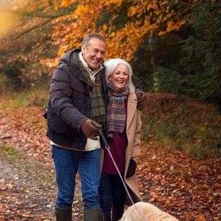 Herbstblues – Symptome und Tipps: Ein Paar geht mit einem Hund durch den herbstlichen Wald spazieren und freut sich.