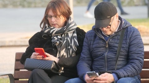 Handysucht: Zwei Menschen sitzten nebeneinander auf einer Bank und schauen auf ihre Handys.