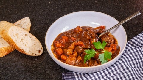 Das ungarische Gulasch wird in einem weißen Teller präsentiert. Neben dem Teller sind zwei aufgeschnittene Baguettscheiben platziert. Auf der anderen Seite des Tellers liegt ein Geschirrtuch.