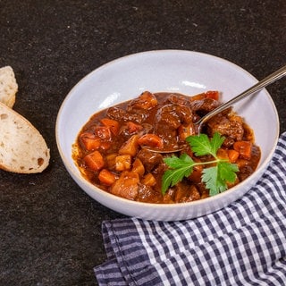 Das ungarische Gulasch wird in einem weißen Teller präsentiert. Neben dem Teller sind zwei aufgeschnittene Baguettscheiben platziert. Auf der anderen Seite des Tellers liegt ein Geschirrtuch.