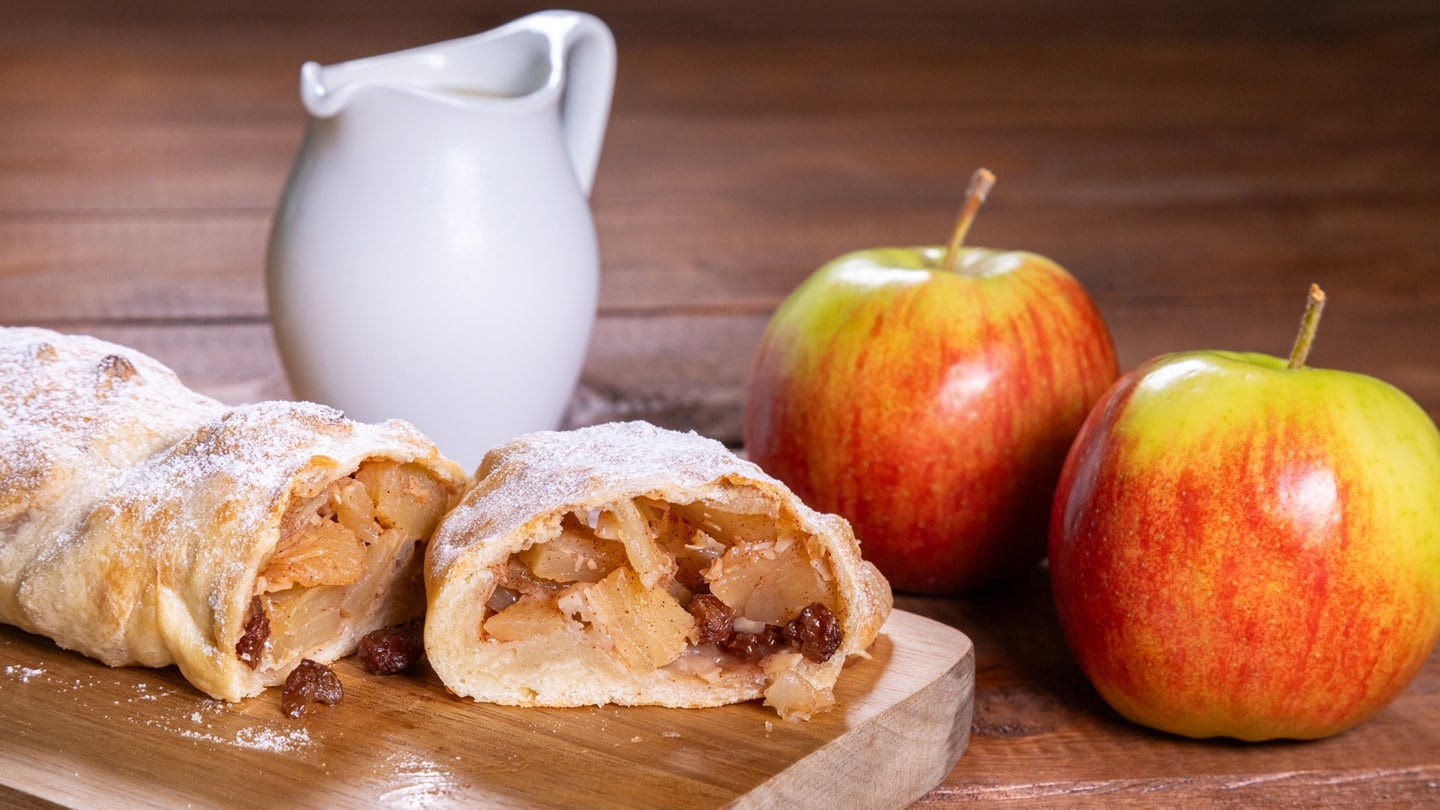 Rezept für Apfelstrudel: Leckerer Apfelstrudel bestäubt mit Puderzucker und Vanillesoße. Der Apfelstrudel ist mit zwei Äpfeln und einem Kännchen mit Vanillesoße angerichtet.