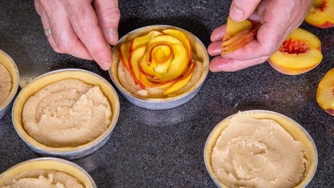 Pfirsichtartelettes backen: Pfirsiche in dünne Scheiben schneiden und zu einer Rose auf dem Mürbeteig garnieren.