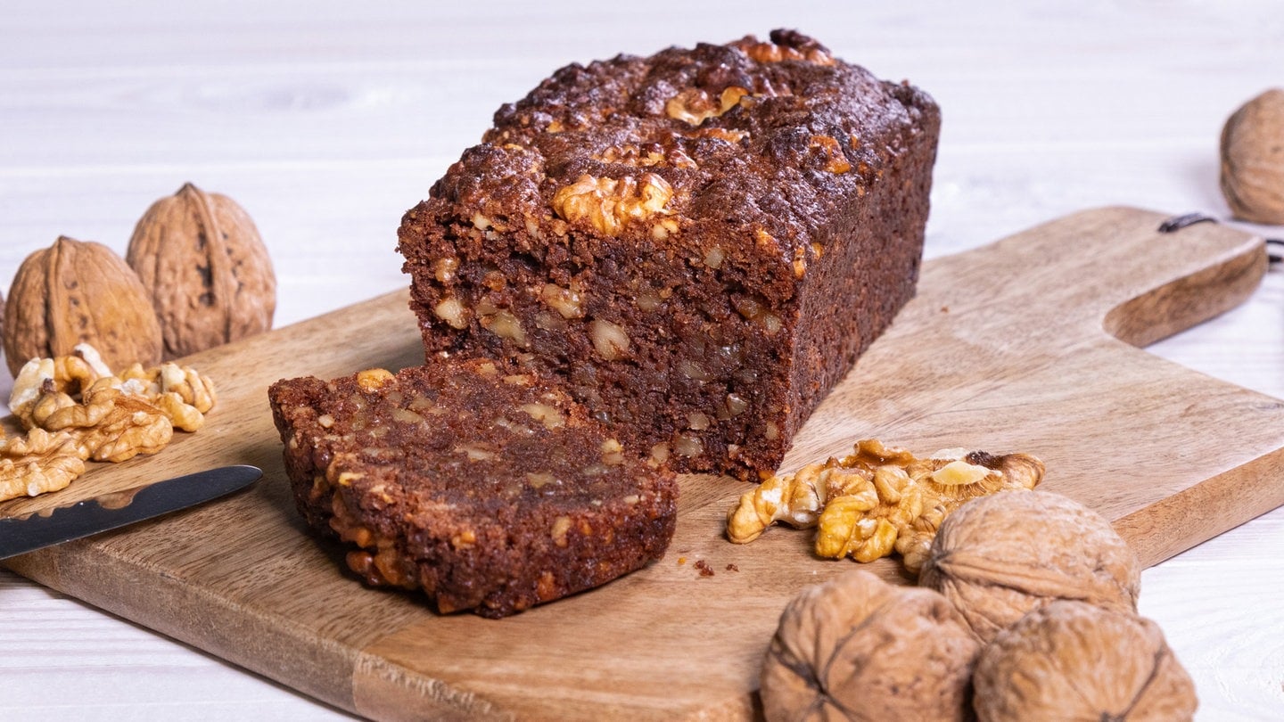 Veganer Kuchen mit Walnuss, der auf einem Holzbrett angerichtet und aufgeschnitten ist. Der saftige Kuchen ist mit ganzen und halben Walnüssen dekoriert.