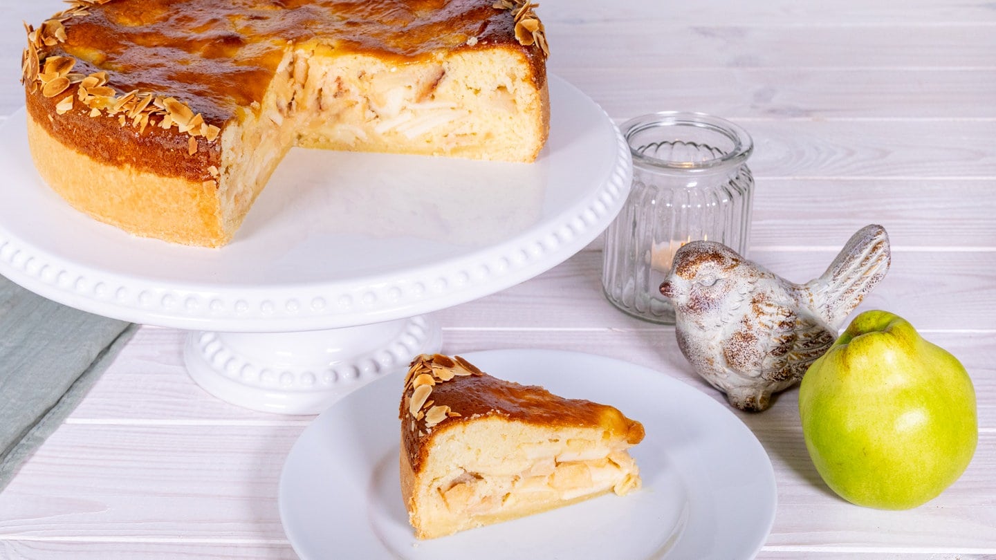 Rezept für einfachen Kuchen aus Quitten und Äpfeln auf Mürbeteig: der fertige Kuchen, angeschnitten auf einer Kuchenplatte, ein Stück auf einem Teller. daneben eine Quitte und ein Deko-Vogel