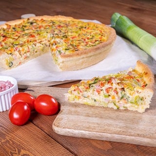 Rezept Lauchkuchen: Der Lauchkuchen ist angerichtet auf einem Holzbrett. Daneben liegt ein Lauch, kleine Tomaten und ein Schälchen mit Schinkenwürfeln.