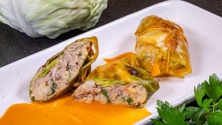 Rezept Kohlrouladen mit Hackfleisch: Die Kohlrouladen sind in der Mitte aufgeschnitten, so dass man die Hackfleischfüllung sehen kann. Das Gericht ist auf einem weißen Teller angerichtet, im Hintergrund liegt ein weißer Kohlkop, im Vordergrund ein Bund frische Petersilie.