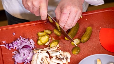 Geschnittene Gurken auf dem Schneidebrett neben Pilzen und Zwiebeln: Für das Bauernfrühstück Hoppelpoppel, wie es in Berlin Tradition hat, dürfen diese Zutaten nicht fehlen.