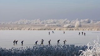 Mehrere Eisläufer fahren auf einem zugefrorenen See in Winterlandschaft Schlittschuhe.