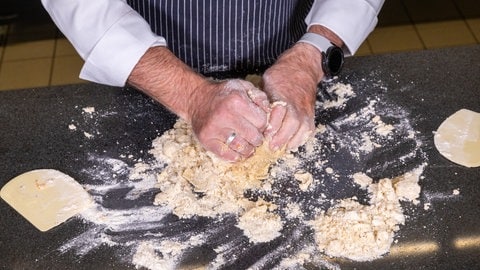 Mürbeteig für den Apfelkuchen mit Gitter: Zwei Hände kneten einen Teig, daneben liegen zwei Teigkarten aus Plastik.