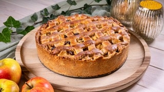 Rezept für Apfelkuchen mit Gitter: Goldbraun gebacken und sehr fein angerichtet steht der saftige Kuche auf einem Tablett. Äpfel liegen daneben.