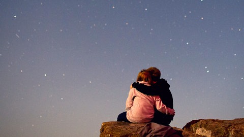 Ein junges Paar betrachtet auf einem Felsen den Sternenhimmel. Sternzeichen, Horoskope und Astrologie im Allgemeinen faszinieren viele Menschen.