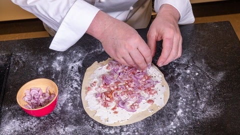 Ein Koch belegt einen original Elsässer Flammkuchen mit Schmand, Zwiebeln und Speck.