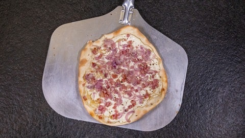 Ein original Elsässer Flammkuchen mit Speck, Zwiebeln und Schmand belegt liegt auf einem Pizzaschieber, der auf einer schwarzen Arbeitsplatte liegt.