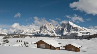 Reise-Doku verschneites Südtirol. Man sieht eine verschneite Alm mit zwei Holzhäusern und im Hintergrund weiße Berge.