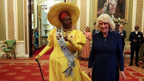 Links geht eine Frau in einem gelben Kostüm mit Hut durch den Buckingham Palace, begleitet von rechts daneben der blonden Königin Camilla in dunklem Kleid. Sie unterhalten sich.