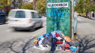 Ein Kleiderberg liegt vor einem Altkleidercontainer mit der Aufschrift «Kleiderbox» am Straßenrand
