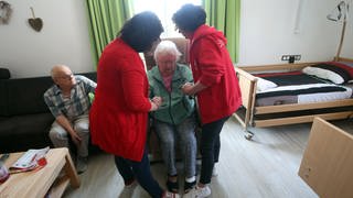 Pflegerinnen helfen pflegebedürftiger Frau aus einem Sessel