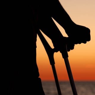 Ein Mann mit zwei Krücken steht bei Sonnenuntergang am Ostseestrand von Warnemünde.